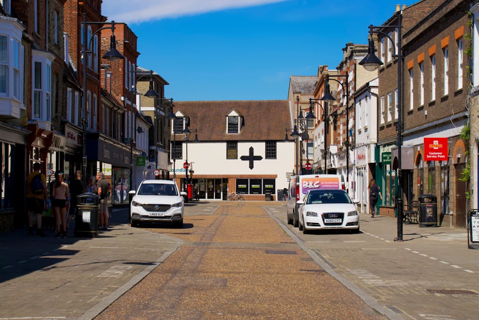 A view of Uxbridge High Street during daytime with a clear blue sky, showing a commercial area with multiple shops and pedestrians on the sidewalks. In the foreground, a white van is parked on the left side, while a silver car and a white Audi are parked on the right side. Both vehicles are positioned near the curb, adjacent to street lamps and a red customer service point sign attached to one of the building facades. Several people are visible walking or standing, some near shop entrances, with a group gathered under the awning of a shop on the left. The street is paved with cobblestone uplighting, and the surroundings include multi-storey brick buildings with various storefronts and signage. The scene depicts an active commercial district with potential considerations for house removals or furniture transport in the area, suitable for logistical planning by [COMPANY_NAME] for relocating items through this part of Uxbridge.
