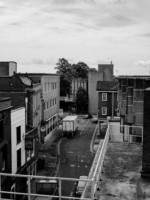 Black and white photograph of a narrow urban street viewed from a higher vantage point, showing various buildings lining both sides, including brick and modern facades with multiple windows. The street is partially blocked by a white delivery truck positioned in the middle of the road, facing away from the camera. To the right, a building with a flat roof and exposed brickwork is visible, while on the left, a building with a sign attached near the corner can be seen. The street has markings for parking and restricted zones, with a few cars visible parked along the curbs. Overhead, a partly cloudy sky is seen, and in the background, trees and additional buildings further down the street are visible. This scene depicts a typical high street environment, relevant for house removals and furniture transport, and illustrates elements involved in the logistics and planning of a home relocation, including street access and vehicle positioning. The image aligns with the services offered by Uxbridge Removals, focusing on urban moving logistics.